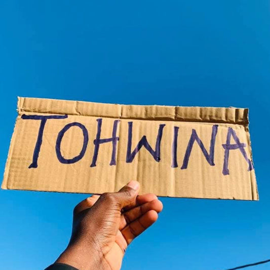 Hand holding a cardboard sign with 'TOHWINA' against a blue sky